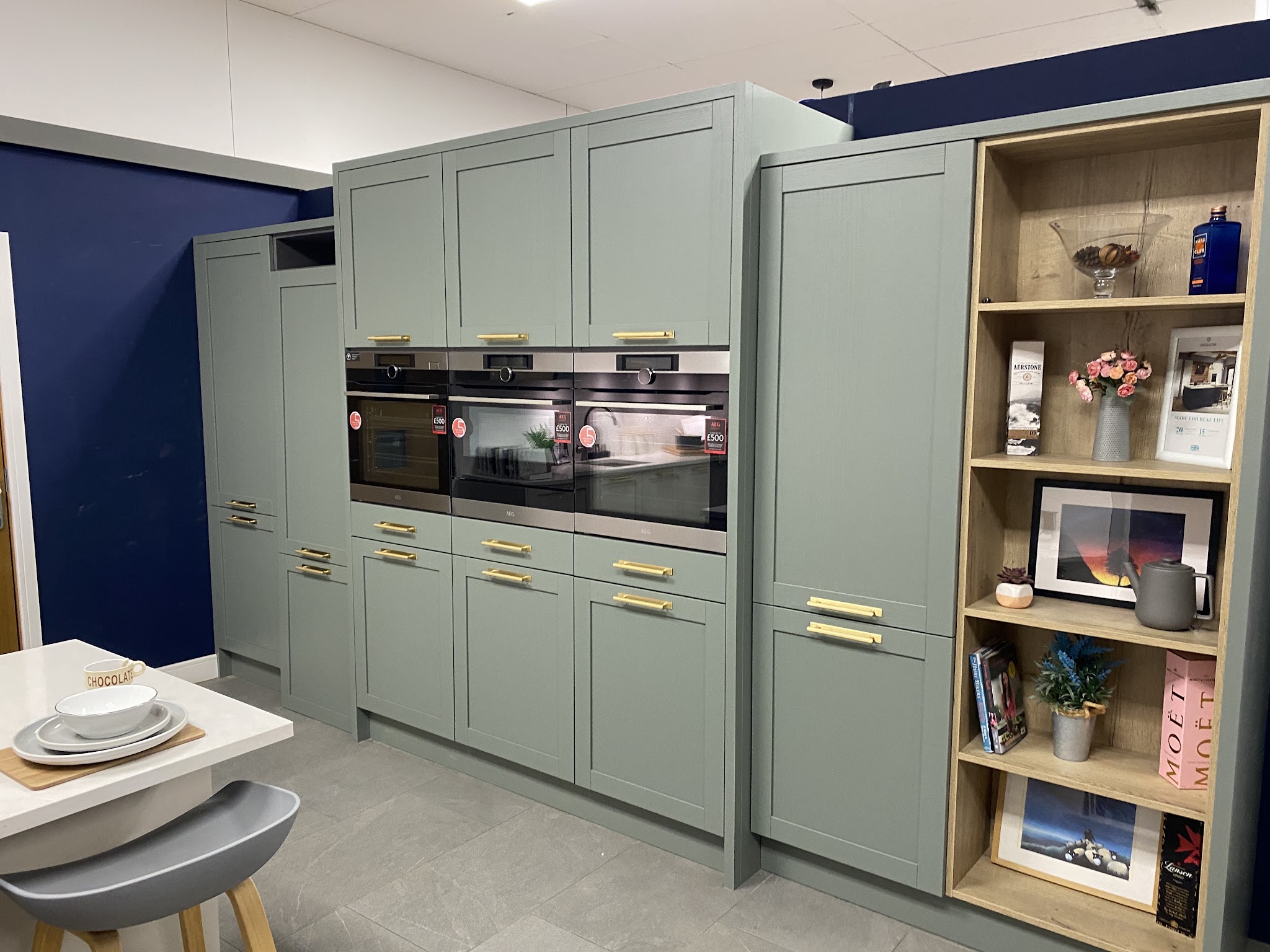 Sage green shaker tall units with brass handles and an oak dresser shelf in a showroom